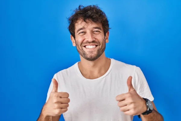 ヒスパニック系の若い男が青の背景の成功のサインの上に立って手で積極的なジェスチャーを行うと 親指を笑顔と幸せ 陽気な表情と勝者のジェスチャー — ストック写真