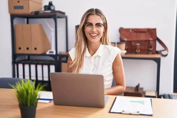 Young blonde woman working at the office wearing glasses looking positive and happy standing and smiling with a confident smile showing teeth