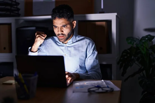 Hispanic man with beard working at the office with laptop at night annoyed and frustrated shouting with anger, yelling crazy with anger and hand raised