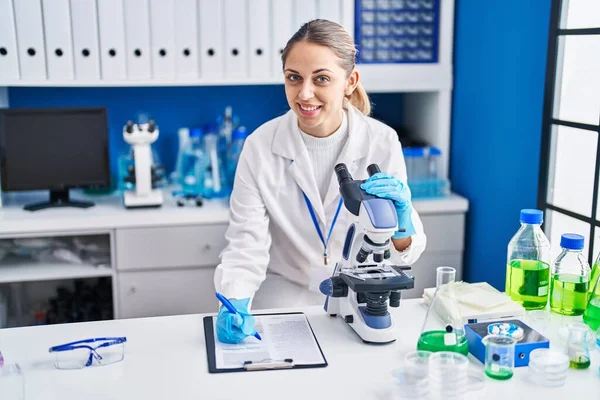 Young woman scientist using microscope writing on report at laboratory