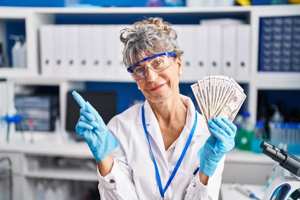 Middle age woman working at scientist laboratory holding dollars smiling happy pointing with hand and finger to the side
