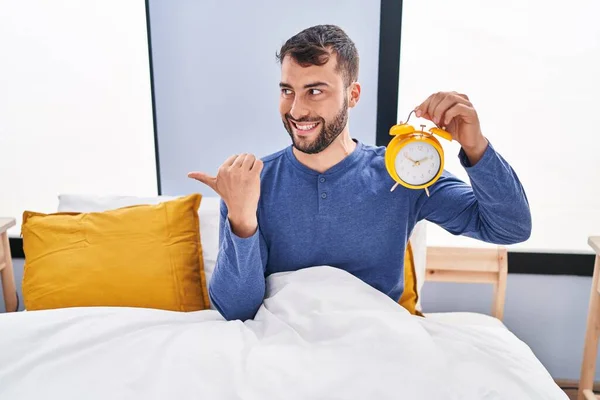 Handsome Hispanic Man Bed Holding Alarm Clock Pointing Thumb Side — Stock fotografie