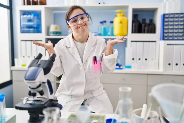 Hispanic girl with down syndrome working at scientist laboratory clueless and confused expression with arms and hands raised. doubt concept.