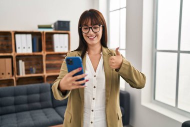 Ofiste çalışan genç esmer kadın akıllı telefonuyla gülümsüyor, mutlu ve pozitif, başparmak yukarı, mükemmel ve onay işareti yapıyor. 