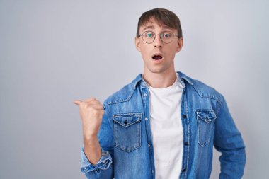 Caucasian blond man standing wearing glasses surprised pointing with hand finger to the side, open mouth amazed expression. 