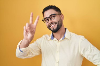 İş kıyafetleri ve gözlük takan İspanyol genç adam kameraya bakıp zafer işareti yapıyor. İki numara.. 