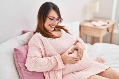 Young pregnant woman doing heart gesture with hands sitting on bed at bedroom