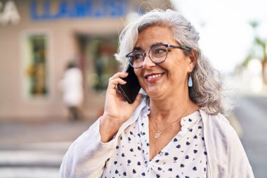 Orta yaşlı bir kadın gülümsüyor. Sokakta akıllı telefondan konuşuyor.