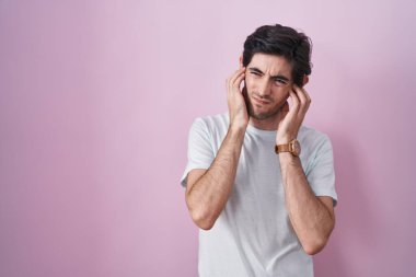 Young hispanic man standing over pink background covering ears with fingers with annoyed expression for the noise of loud music. deaf concept. 