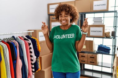 Young african american woman wearing volunteer t shirt at donations stand gesturing finger crossed smiling with hope and eyes closed. luck and superstitious concept. 