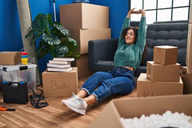 Genç, güzel, İspanyol bir kadın yeni evinde yerde oturmuş kollarını açıyor.