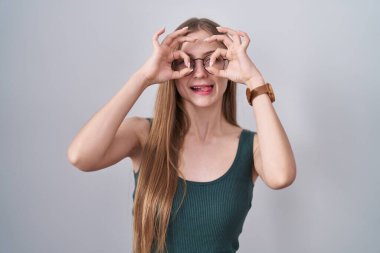 Young caucasian woman standing over white background doing ok gesture like binoculars sticking tongue out, eyes looking through fingers. crazy expression. 