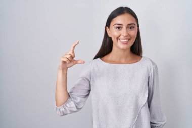 Young hispanic woman standing over white background smiling and confident gesturing with hand doing small size sign with fingers looking and the camera. measure concept. 
