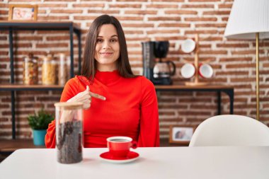 Young hispanic woman drinking coffee at home pointing finger to one self smiling happy and proud 