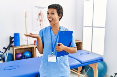 Kısa saçlı, kısa saçlı bir İspanyol kadın ağrı kesici kliniğinde çalışıyor. Avuç içleri açık, fotokopi alanını gösteriyor. Gülümseyen mutlu bir gülümseme var. 