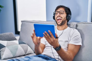 Young hispanic man watching video on touchpad sitting on sofa at home