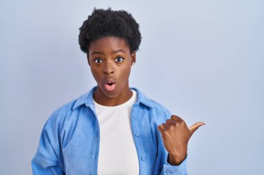 Mavi arka planda dikilen Afro-Amerikan kadın, elleri yana doğru işaret ederek şaşırdı, ağzı açık hayret içinde kaldı.. 