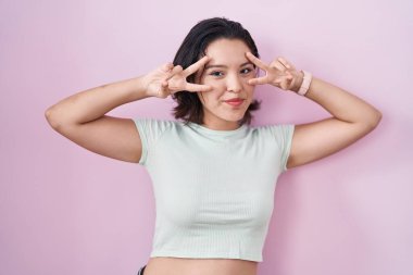 Hispanic young woman standing over pink background doing peace symbol with fingers over face, smiling cheerful showing victory 