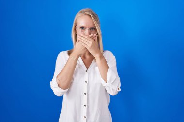 Mavi arka planda duran genç beyaz kadın yanlışlıkla elleri ile ağzını kapatırken şok oldu. gizli kavram. 