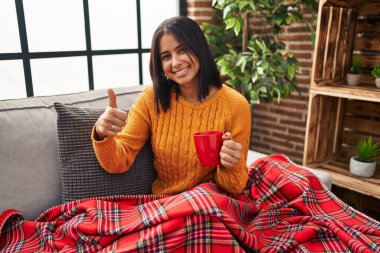 Kanepede oturan Latin asıllı genç bir kadın evde kahve içerek olumlu bir jest yapmayı onaylıyor, gülümsüyor ve başarı için seviniyor. kazanan jest. 