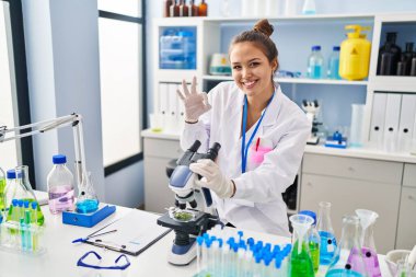 Laboratuvarda çalışan Latin asıllı genç bir kadın parmaklarıyla imza atıyor, gülümseyerek mükemmel bir sembol oluşturuyor. 