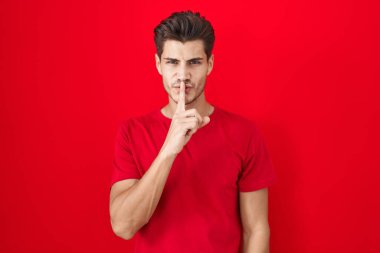 Genç İspanyol adam kırmızı arka planda duruyor ve parmakları dudaklarında sessiz olmak istiyor. sessizlik ve gizli konsept. 