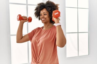 Genç Afrikalı Amerikalı kadın gülümsüyor spor merkezinde halterleri kullanarak kendine güven eğitimi alıyor