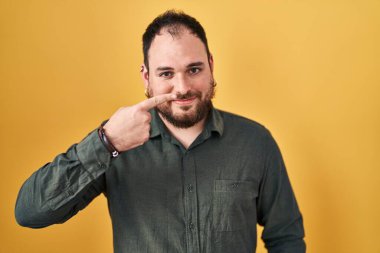 Plus size hispanic man with beard standing over yellow background pointing with hand finger to face and nose, smiling cheerful. beauty concept 