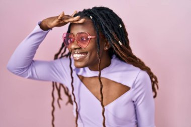 African woman with braided hair standing over pink background very happy and smiling looking far away with hand over head. searching concept. 