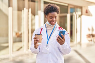 Doktor üniforması ve tıbbi maske takan Afro-Amerikalı kadın akıllı telefon kullanarak sokakta kahve içiyor.