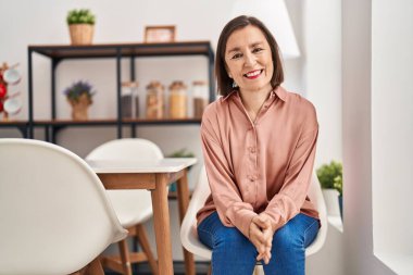 Orta yaşlı bir kadın evdeki sandalyede otururken kendinden emin gülümsüyor.