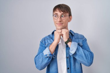 Caucasian blond man standing wearing glasses laughing nervous and excited with hands on chin looking to the side 