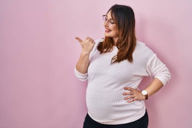 Pregnant woman standing over pink background smiling with happy face looking and pointing to the side with thumb up. 