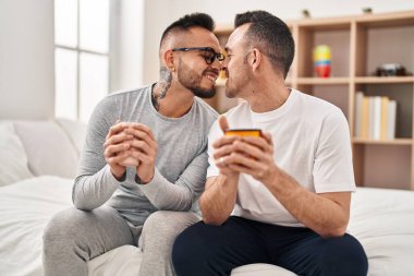 Two men couple drinking cup of coffee sitting on bed at bedroom