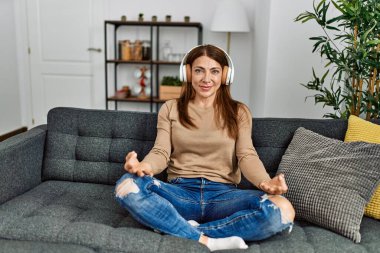 Orta yaşlı beyaz kadın yoga yapıyor ve kulaklık takıyor. Evdeki kanepede oturuyor..