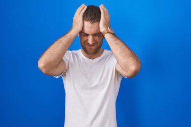 Young caucasian man standing over blue background suffering from headache desperate and stressed because pain and migraine. hands on head. 