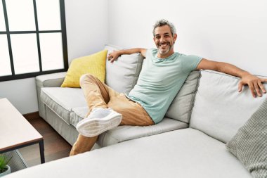 Orta yaşlı, kır saçlı, gülümseyen bir adam. Evdeki kanepede oturuyor.