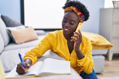 Young african american woman writing on notebook talking on the smartphone at home