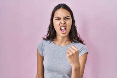 Genç Brezilyalı bir kadın pembe arka plan üzerine gündelik tişört giyip öfkeyle bağırırken kızgın ve sinirli bir şekilde yumruğunu kaldırıyor. öfke ve agresif konsept. 