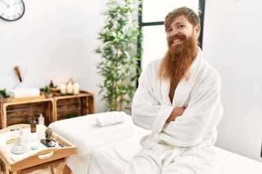 Young redhead man wearing bathrobe sitting on massage table at beauty center