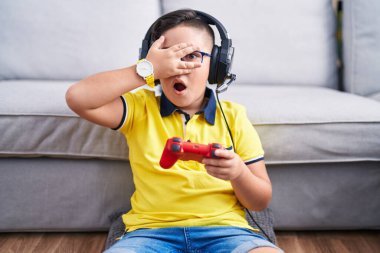 Video oyunu oynayan, kulaklık takan, şok geçirip yüzünü ve gözlerini kapayan utangaç ifadesiyle parmaklarından içeri bakan İspanyol bir çocuk.. 
