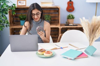Young hispanic girl eating doughnut studying at home