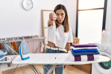Young hispanic woman ironing clothes at laundry room looking unhappy and angry showing rejection and negative with thumbs down gesture. bad expression. 