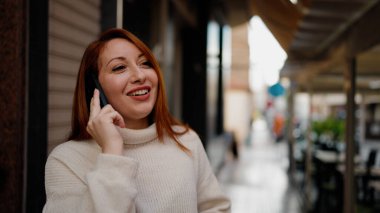 Genç kızıl saçlı, gülümseyen, kendine güvenen, caddedeki akıllı telefondan konuşan kadın.