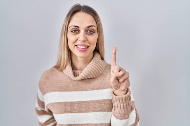 Sarışın genç bir kadın, izole edilmiş arka planda balıkçı yaka kazak giyiyor. Bir numaralı parmağı gösteriyor ve gülümsüyor. Kendinden emin ve mutlu.. 