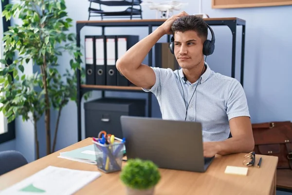 Young hispanic man working at the office wearing headphones confuse and wonder about question. uncertain with doubt, thinking with hand on head. pensive concept.
