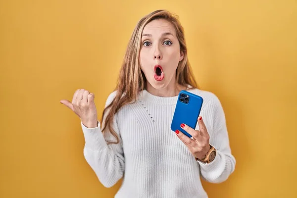 Młoda Blondynka Pomocą Smartfona Wpisując Wiadomość Zaskoczony Wskazując Palcem Bok — Zdjęcie stockowe