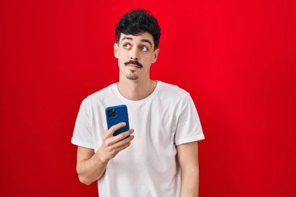 Hispanic Man Using Smartphone Red Background Smiling Looking Side Staring — 图库照片