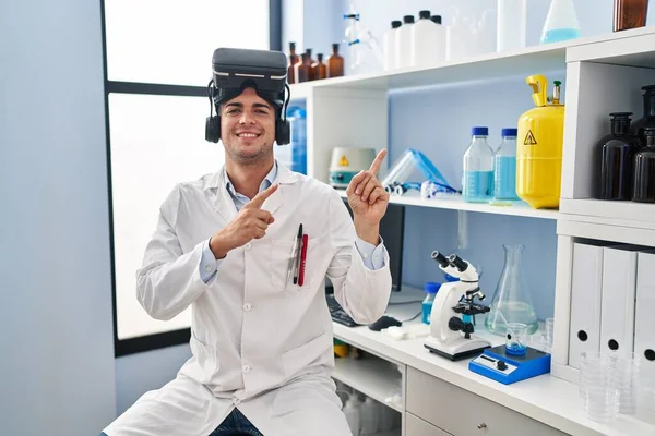 Vrガラスを身に着けている科学研究所で働いている若いヒスパニック系の男は笑顔とカメラを見て2本の手と指で側面を指して — ストック写真