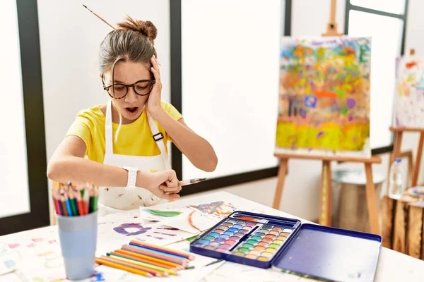 Young brunette teenager at art studio looking at the watch time worried, afraid of getting late